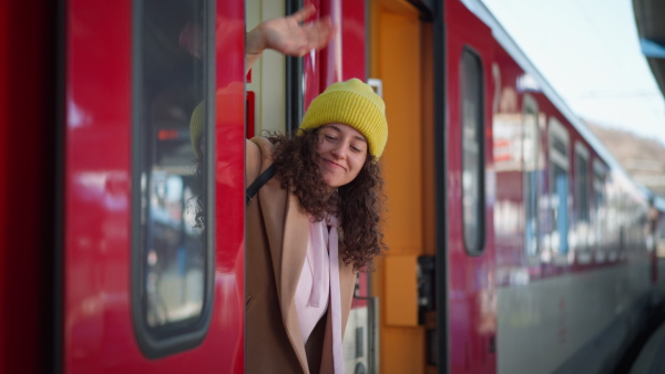 A happy young traveler woman with luggage standing in train door waveing out of train at train station platform