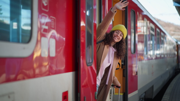 A happy young traveler woman with luggage standing in train door waveing out of train at train station platform