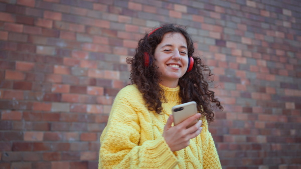 A happy young woman in front of brick wall in city street, looking at camera., listening to music and dancing.