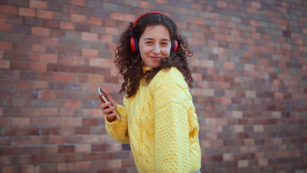 A happy young woman in front of brick wall in city street, looking at camera., listening to music and dancing.