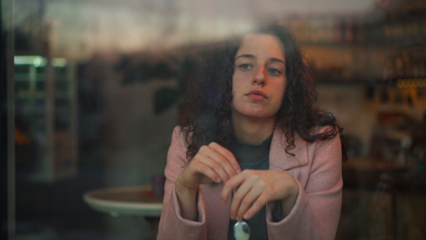 A sad, pensive young woman drinking coffee and looking out of the cafe window.