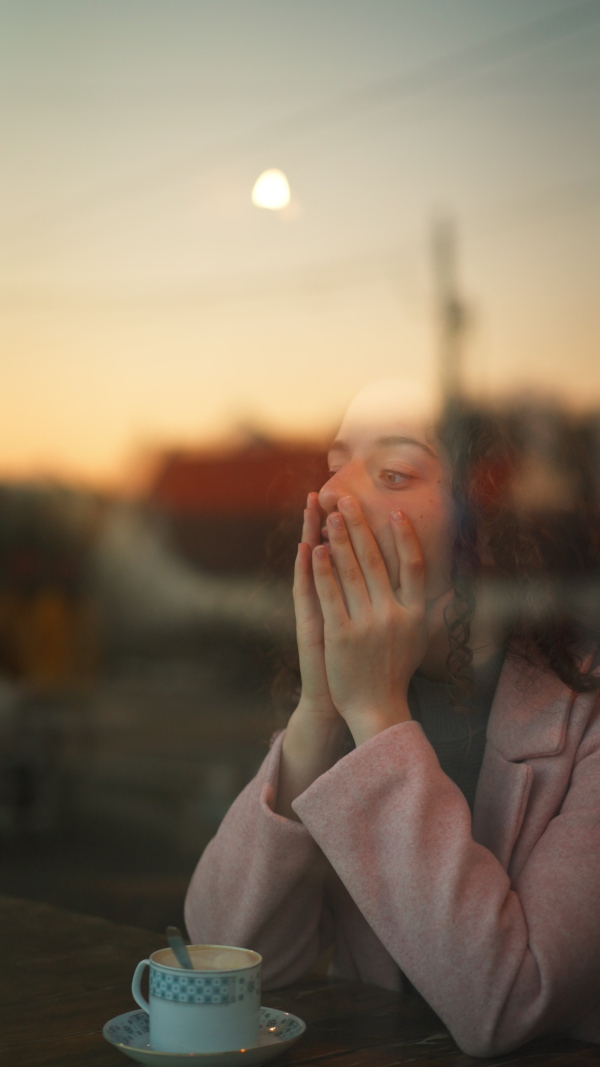 A vertical footage of sad,pensive young woman drinking coffee and looking out of the cafe window.