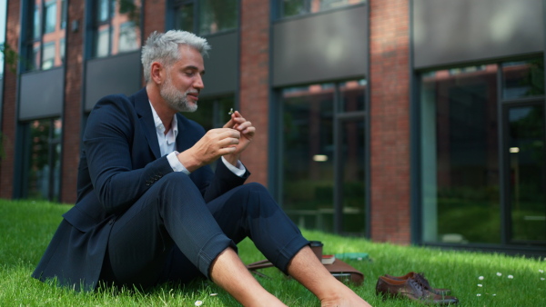 A mature businessman resting and sitting barefoot in park, feeling free, escaping from work, work life balance concept.