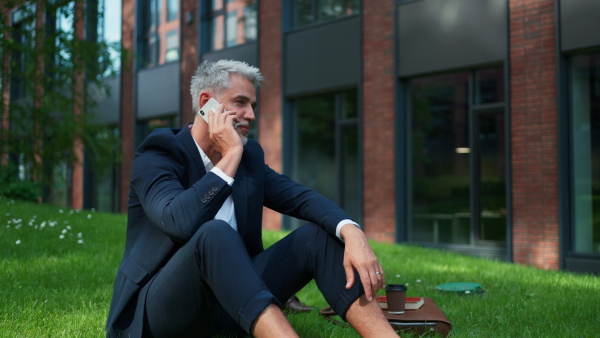 A mature businessman resting and sitting barefoot in park, calling on cellphone, work life balance concept.