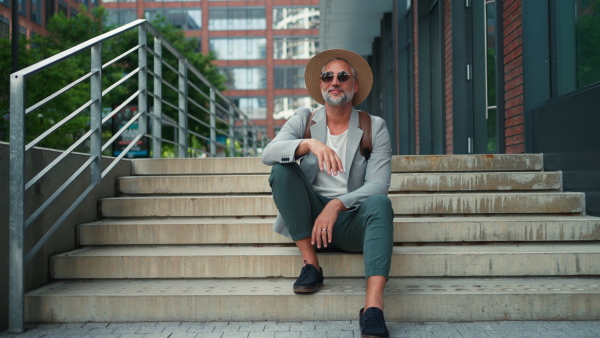 A confident man wearing straw hat and backpack sitting on stairs, businessman in casual clothes in summer on the way to work.