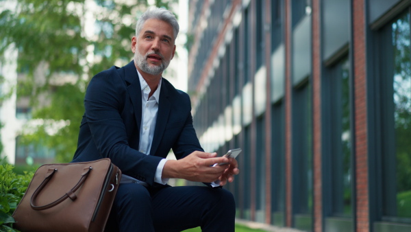 A successful mature businessman using smartphone while resting on street, sitting on bench and using mobile application.