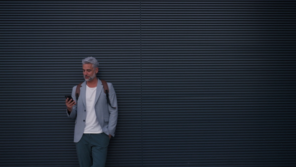 A successful happy businessman waiting in city street, using mobile phone, commuting concept.