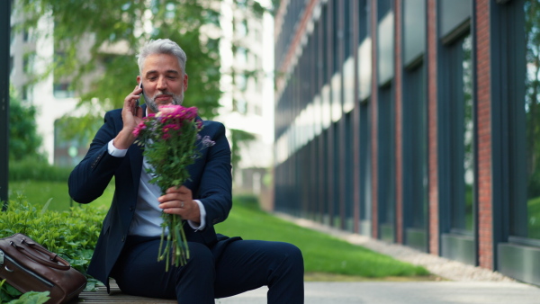 A portrait of happy mature businessman with flower bouquet sitting on bench and waiting for girlfriend.