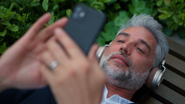 A mature businessman enjoying music in headphones relaxing on bench in city park during break at work.