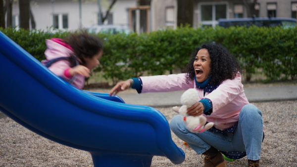 Happy mother playing with child at playground slide. Girl sliding into her mothers arms.