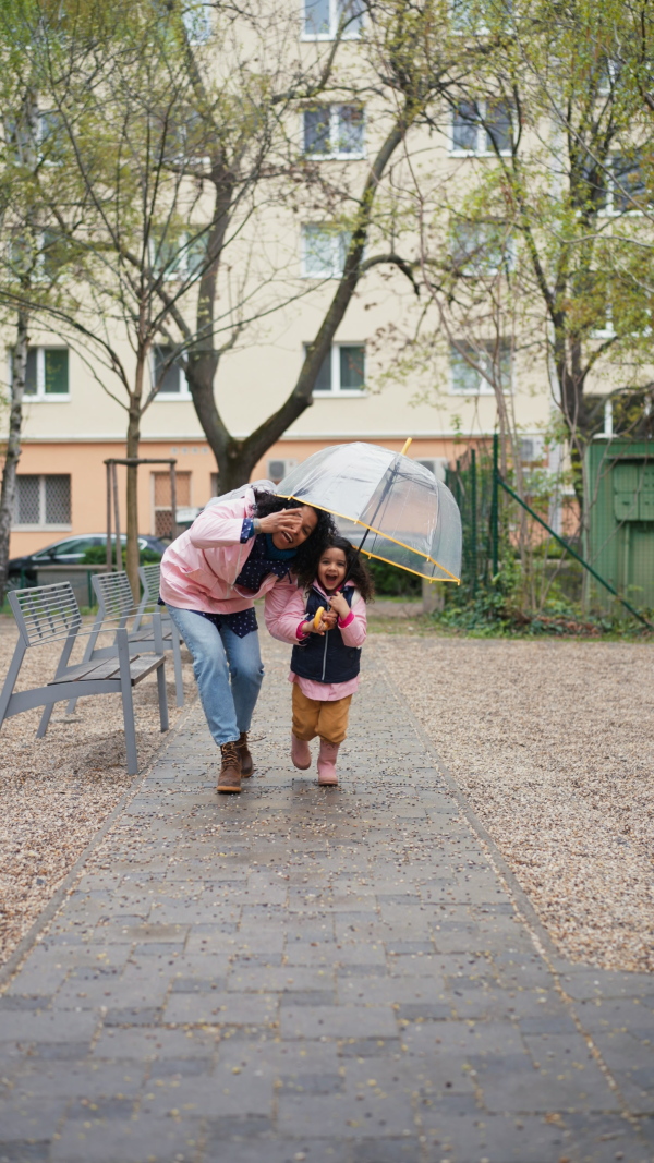 Mother with little daughter runing under an umbrella on rainy day in park, looking into camera. Vertical shot.