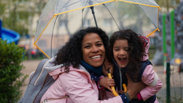 Portrait of smilimg african-american mother and little daughter in rainy weather, hiding under umbrella, looking into camera.