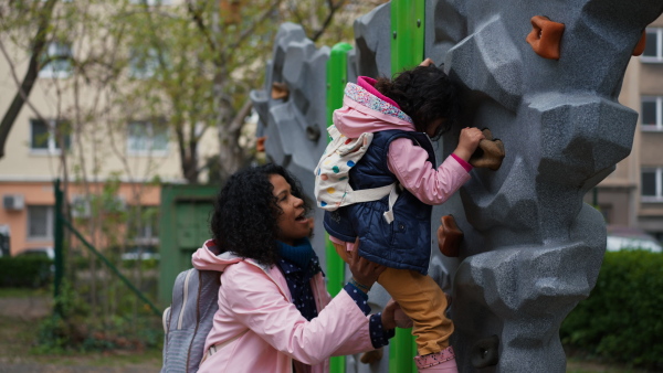 Mother helping little daughter to climb on playground climbing wall.