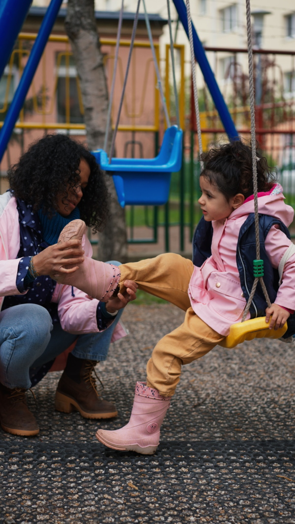 Mother putting on wellington boots to daughter on playground swing in park. Vertical take.