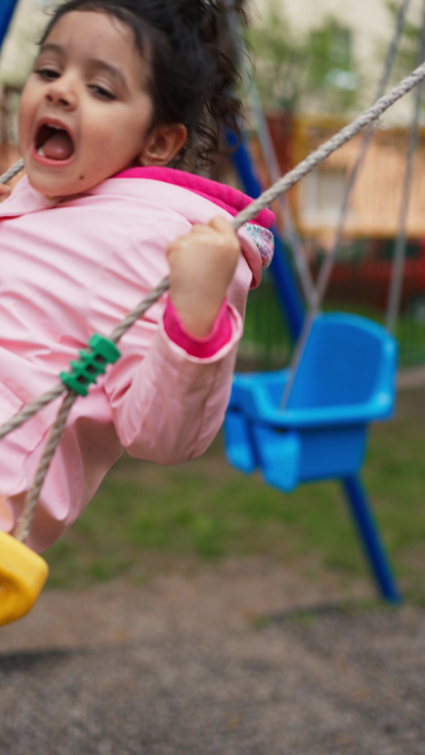 Little girl in pink raincoat laughing on playground swing, having a good time. Vertical shot.