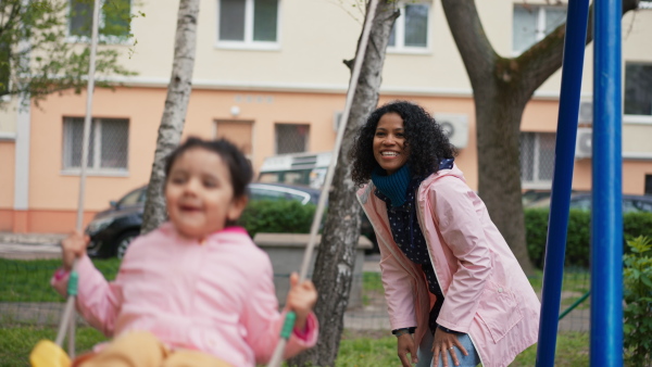 Mother swinging daughter on playground swing in a park, smiling.