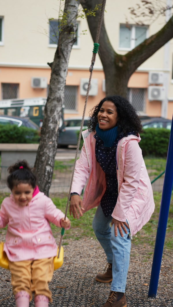 Mother swinging daughter on playground swingin park, smiling.