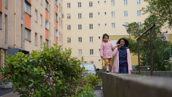 Mother holding hand of little daughter walking on stone wall looking into camera. Apartment buildings and trees in background. Full lenghth shot.