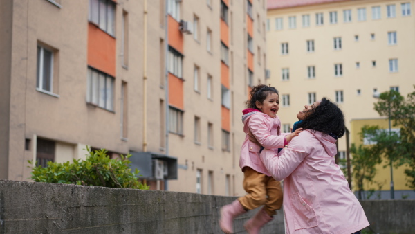 Little girl jumping into mothers arms from small wall in front of apartment building and embracing each other.