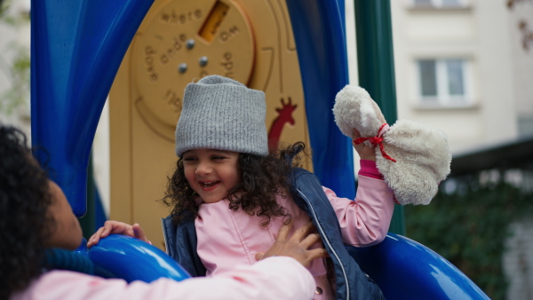 Happy girl on playground slide enjoying to play with her mother, holding a teddybear, smiling.