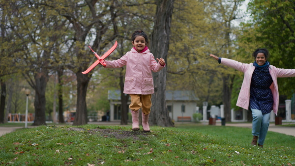 Mother and daughter playing in park with airplane toy, running and imitating flying.