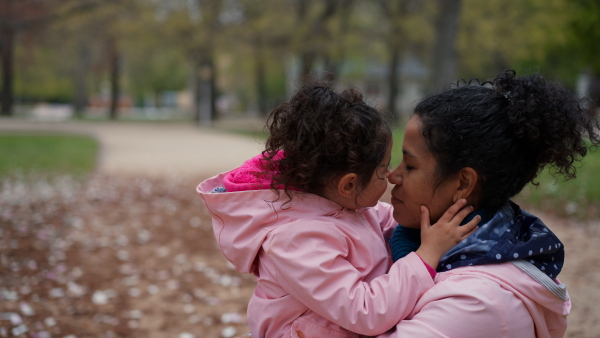 Mother holding little daughter in her arms in park, hugging her tenderly.