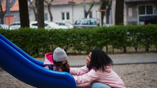 Happy mother playing with child at playground slide. Girl sliding into her mothers arms.