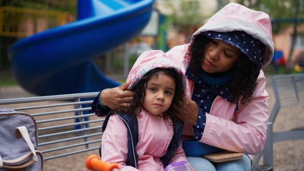 Mother pulling up hood to little daughter on playground in rainy weather. Girl looking into camera.