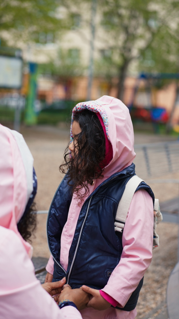 Mother zipping up jacket of little daughter outdoors in park in rainy weather. Vertical shot.