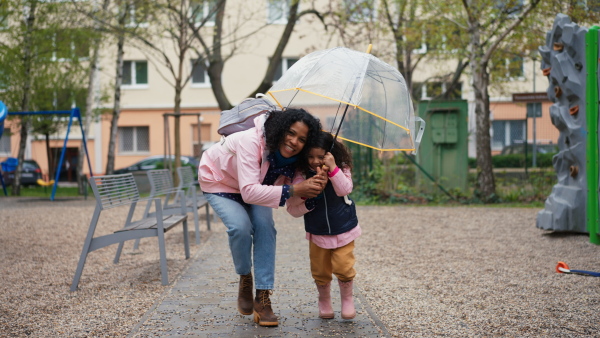 Mother with little daughter runing under an umbrella on rainy day in park, looking into camera.