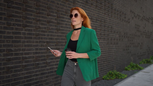 Young woman walking in the city with cup of coffee, scrolling her smartphone.