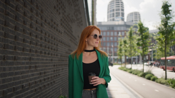 Young stylish redhead woman standing in the city with a sunglasses and cup of coffee.