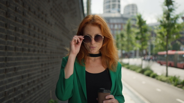 Young stylish redhead woman standing in the city with a sunglasses and cup of coffee.