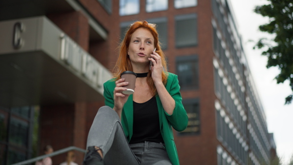 Young woman with cup of coffee sitting in front of the office building and calling.