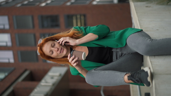 Young woman with cup of coffee sitting in front of the office building and calling.