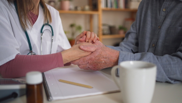 A close-up of doctor holding hand of senior patient and consoling him during medical visit at home.