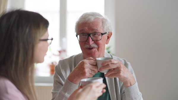 An adult daughter visiting her senior father at home and having coffee together, talking.