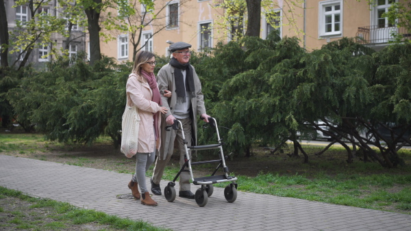 A senior man with walking frame and adult daughter outdoors on a walk in park.