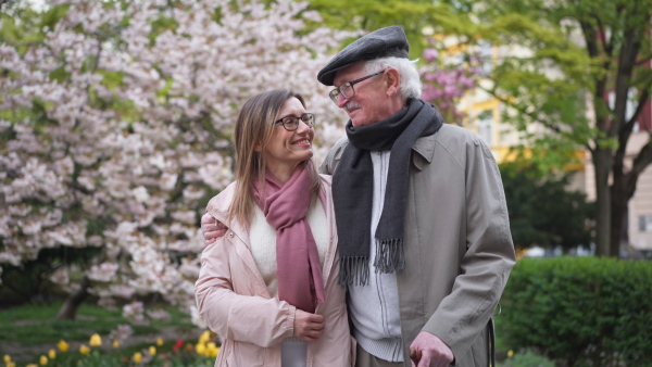 An adult daughter hugging her senior father outdoors in park on spring day.