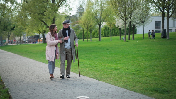 A happy senior man with walking stick and adult daughter outdoors on a walk in park.