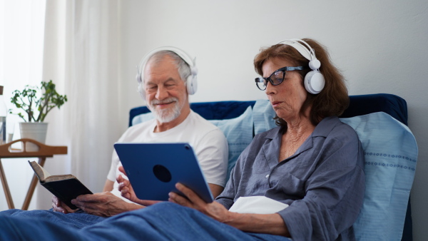 Senior couple in bed, reading book and watching movie on digital tablet.