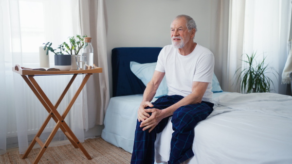 Senior man having pain in his knee, sitting in pyjama on bed.