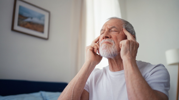 Senior man having headache, sitting in pyjama on bed.