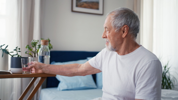 Senior man drinking water in the morning in his bedroom, sitting on bed.