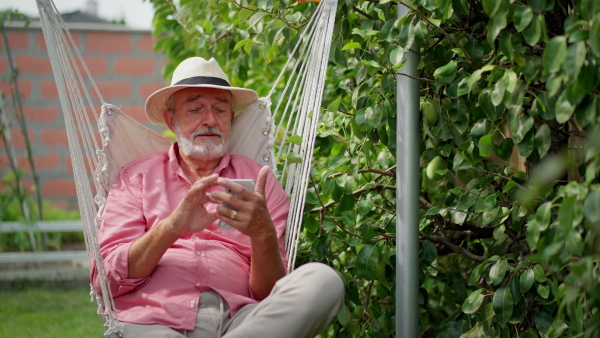 Elederly man sitting relaxed in garden swing, looking at mobile phone.