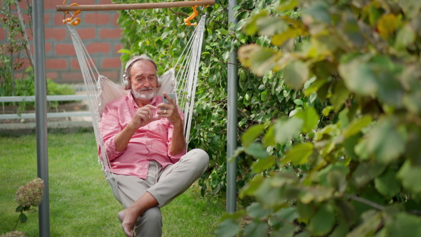 Elderly man sitting on garden swing, listening to music on smart phone, relaxing.