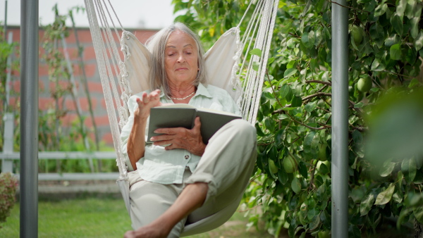 Elderly woman sitting and relaxing in garden swing, reading a book.