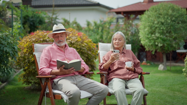 Senior couple sitting in garden, relaxing. Man reading book, woman dinking coffee.