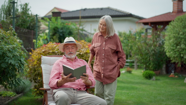 Senior woman approaching husband reading a book, embracing him and smiling.