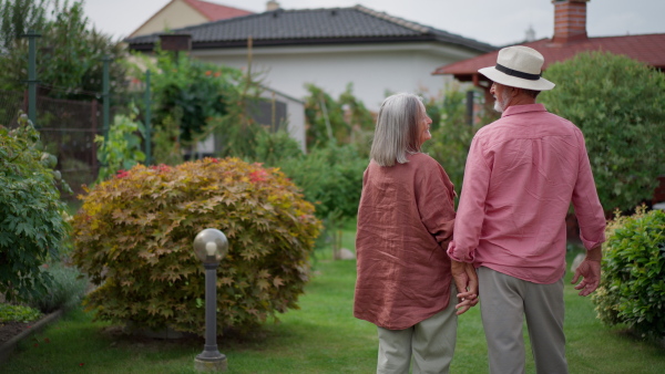 Elderly couple standing in garden, looking at house, holding hands.